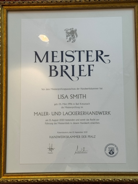 Ein moderner Besprechungsraum mit einem hölzernen runden Tisch und vier Stühlen. An den Wänden hängen mehrere farbige Platten oder Musterstücke in verschiedenen Farbtönen. Drei Pendelleuchten hängen über dem Tisch. Auf dem Tisch steht eine kleine Vase mit Blumen und ein Stapel Papier oder Broschüren. Der Raum hat eine gemütliche und einladende Atmosphäre.